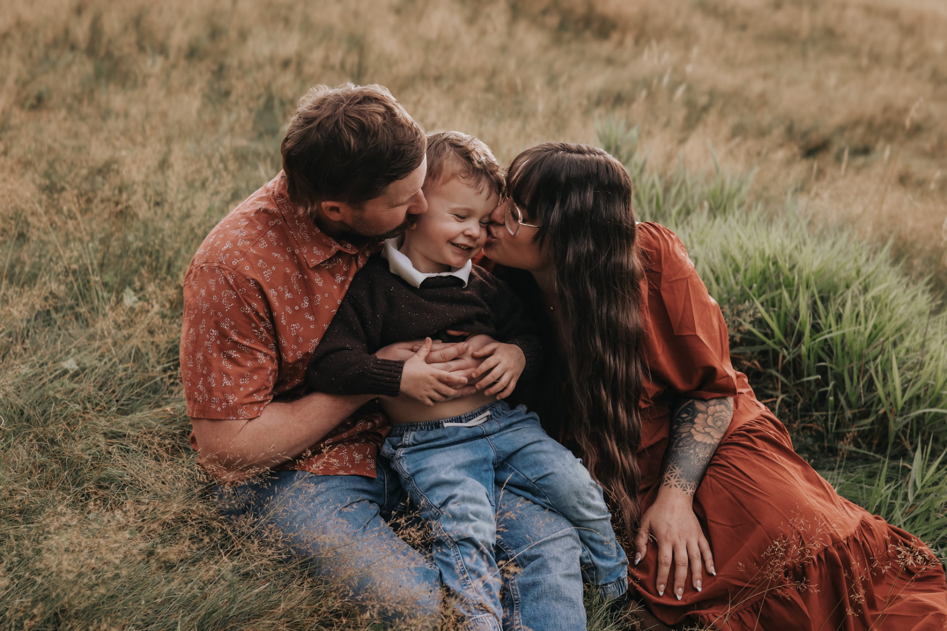 A family sitting in a golden field at sunset — parents kissing their smiling young son on each cheek